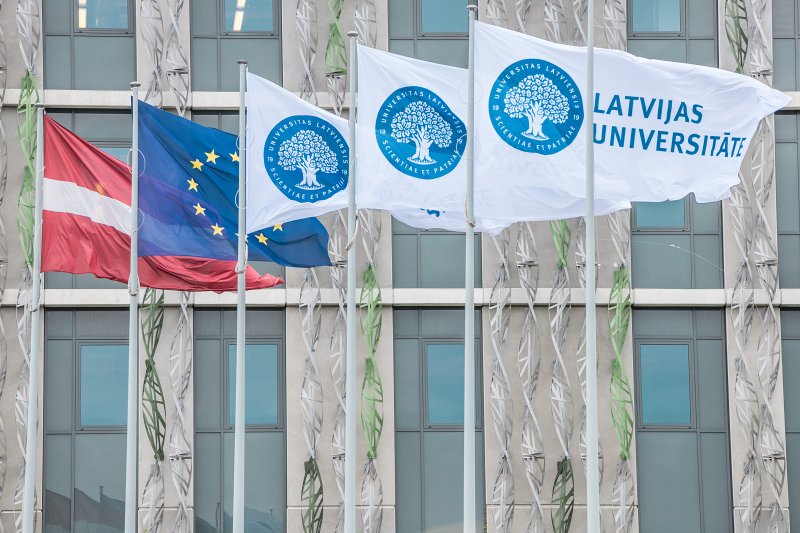 Pie Latvijas Universitātes ēkas fasādes vējā plīvo vairāki karogi: Latvijas karogs, Eiropas Savienības karogs un trīs balti Latvijas Universitātes karogi ar universitātes ģerboni un uzrakstu "Latvijas Universitāte"