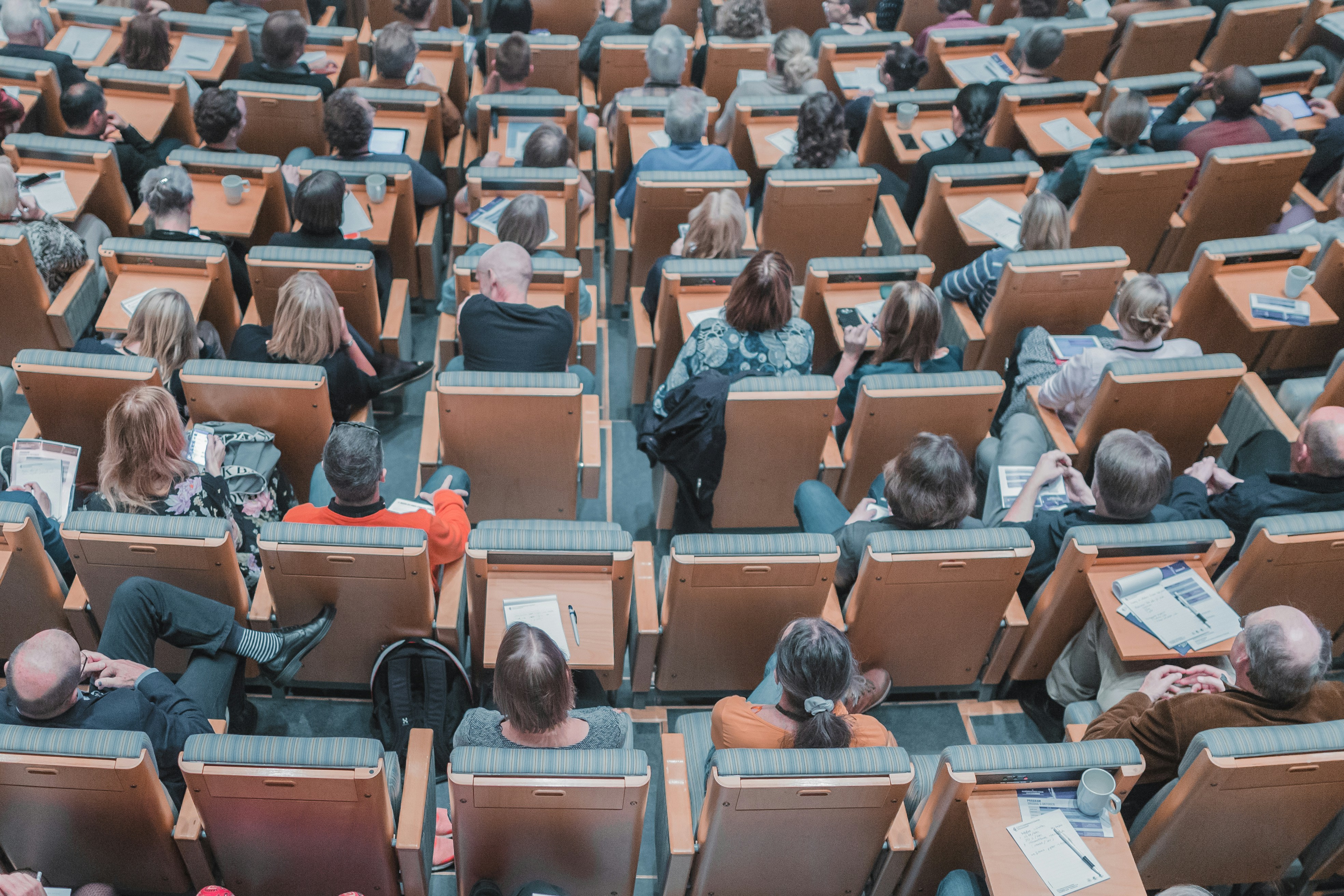 Skats no augšas uz pilnu universitātes auditoriju — daudzi klausītāji sēž koka krēslos ar rakstāmvirsmām un klausās lekciju.