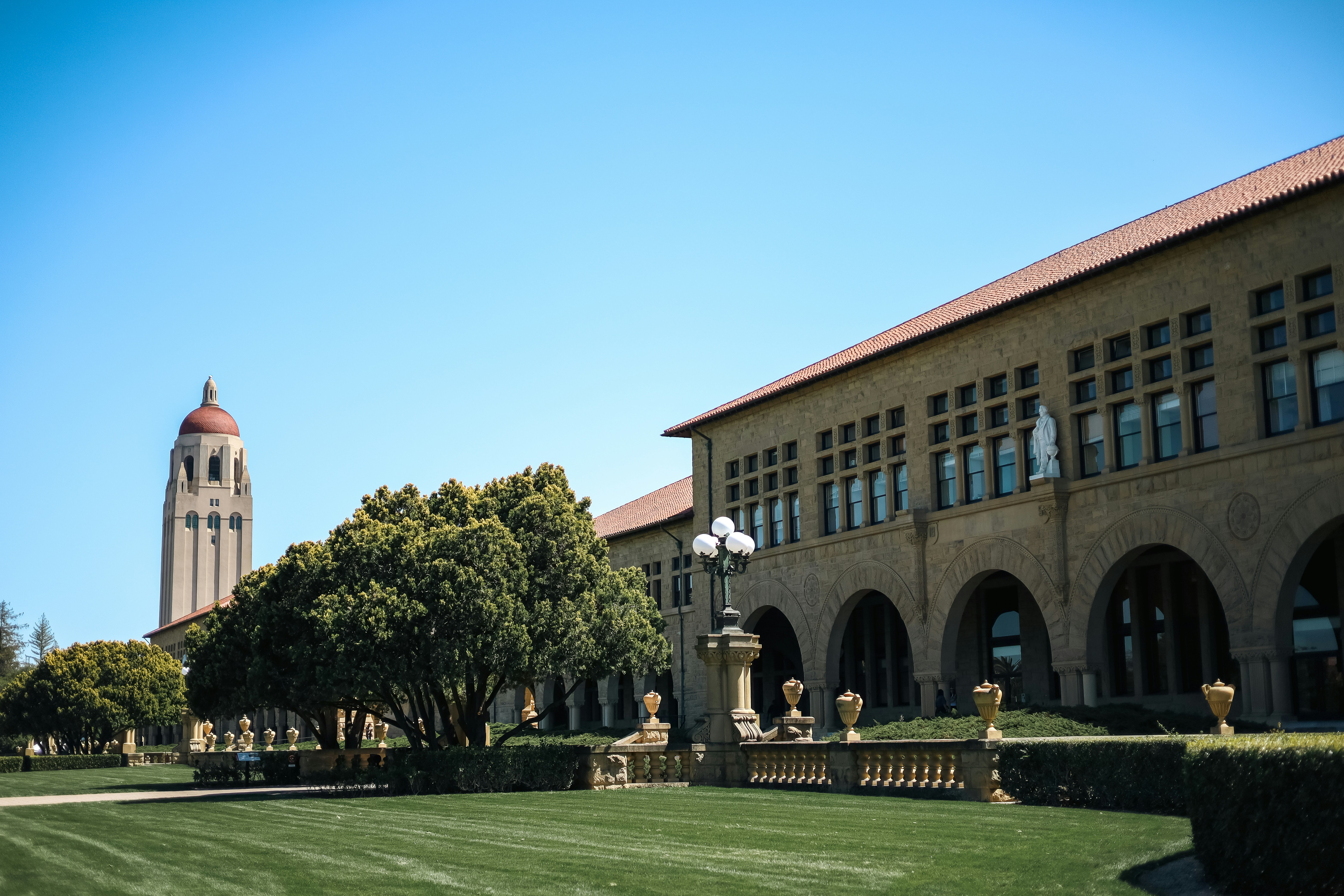 Exterior view of a university campus featuring a historic sandstone building with arched galleries and a red-tiled roof. In the background stands a tall tower with a dome — Hoover Tower at Stanford University, USA.