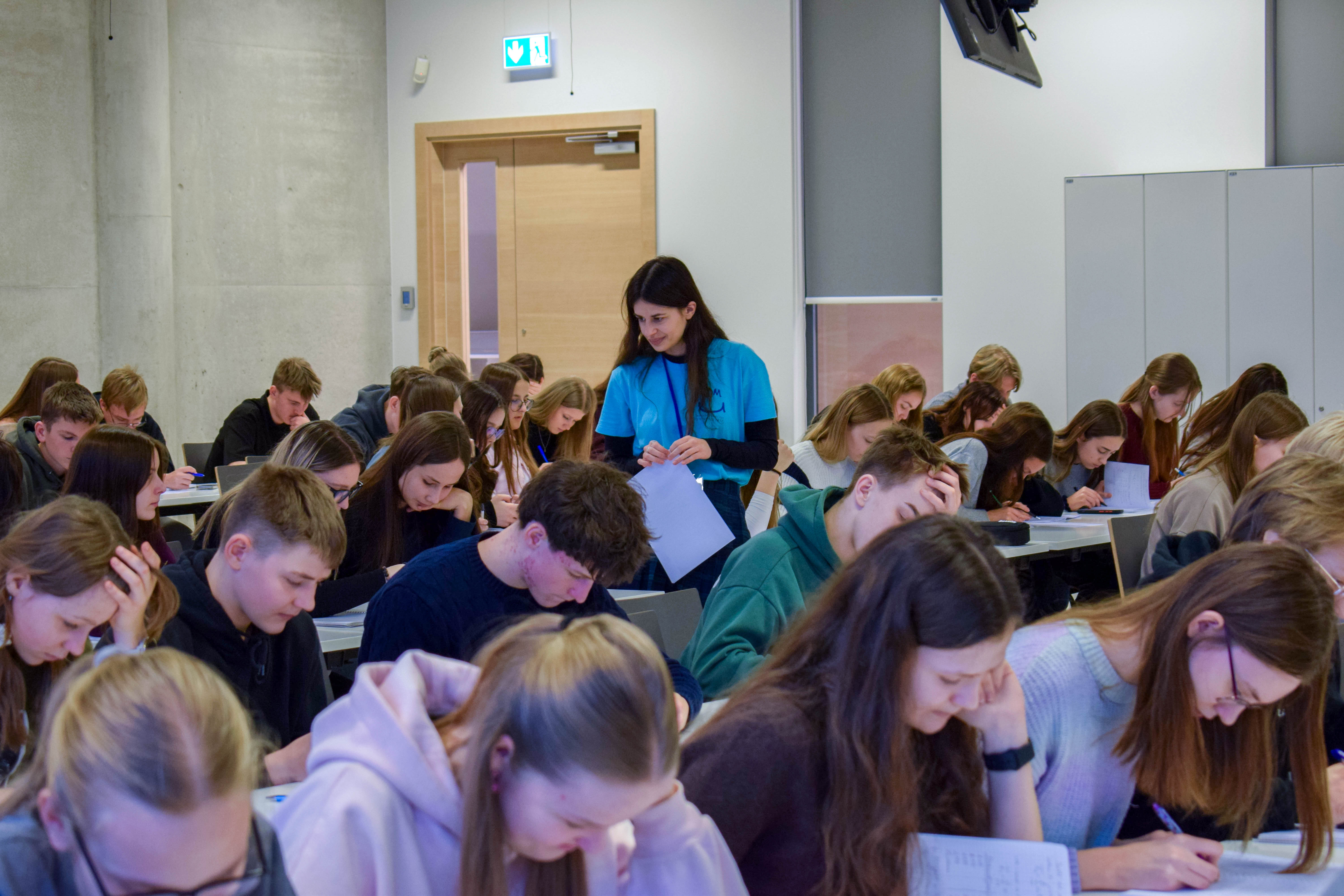 Studente palīdz skolēniem risināt uzdevumus staigājot pa auditoriju