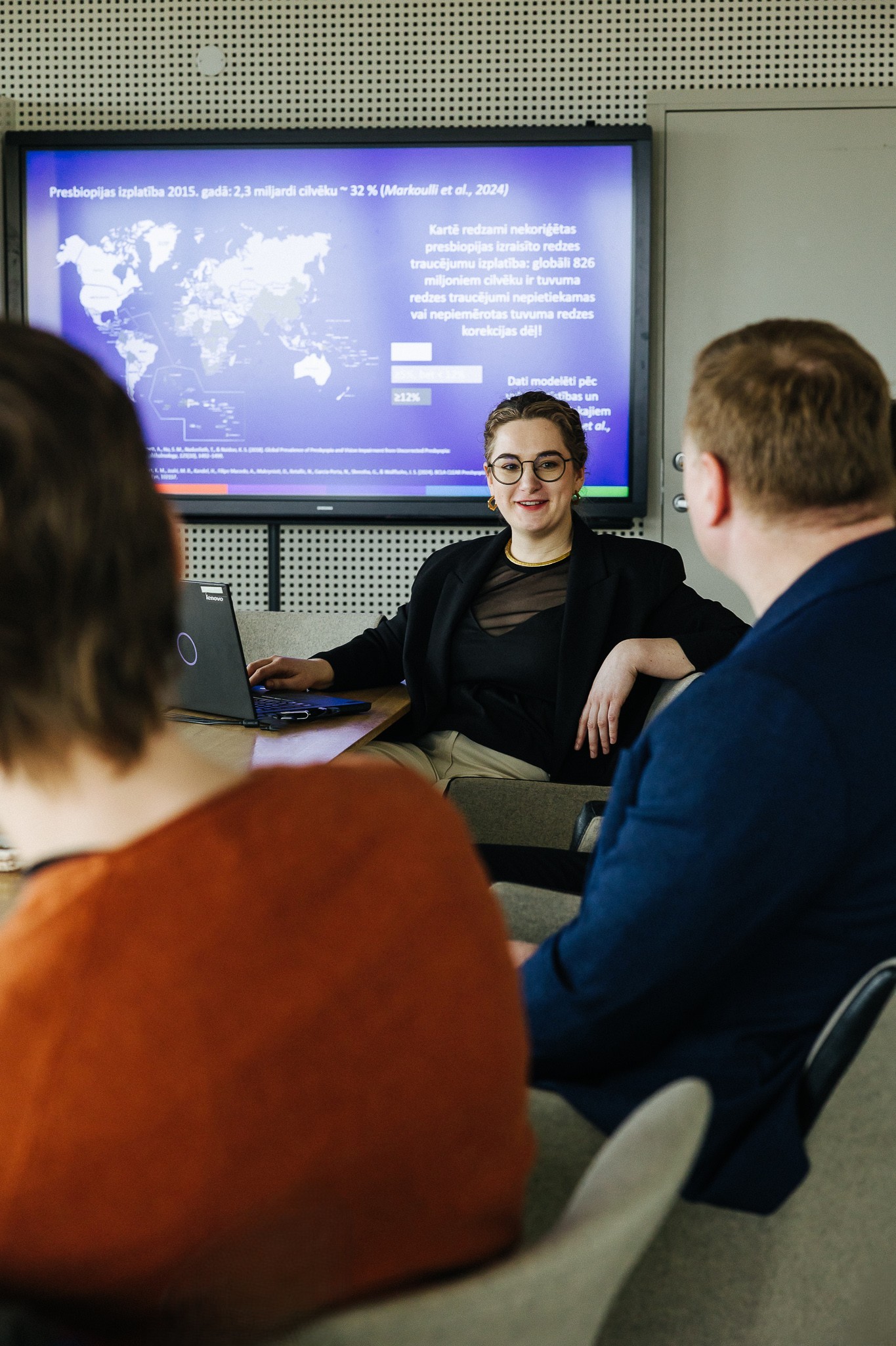 Karola Panke prezentē globālos presbiopijas rādītājus zinātniskajai komandai; slaidā redzama pasaules karte un stabiņu diagramma, kas attēlo nekoriģētas tuvredzes traucējumu izplatību.
