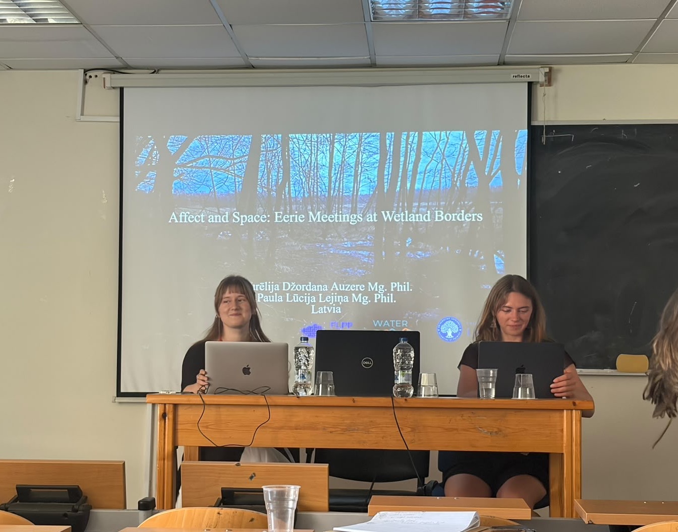 Paula Lūcija Lejiņa and Aurēlija Džordana Auzere sit at a table with their laptops during a conference presentation. Paula Lūcija Lejiņa is on the left and Aurēlija Džordana Auzere on the right. Behind them, the opening slide reads “Affect and Space: Eerie Meetings at Wetland Borders” over a photograph of a wooded wetland landscape.