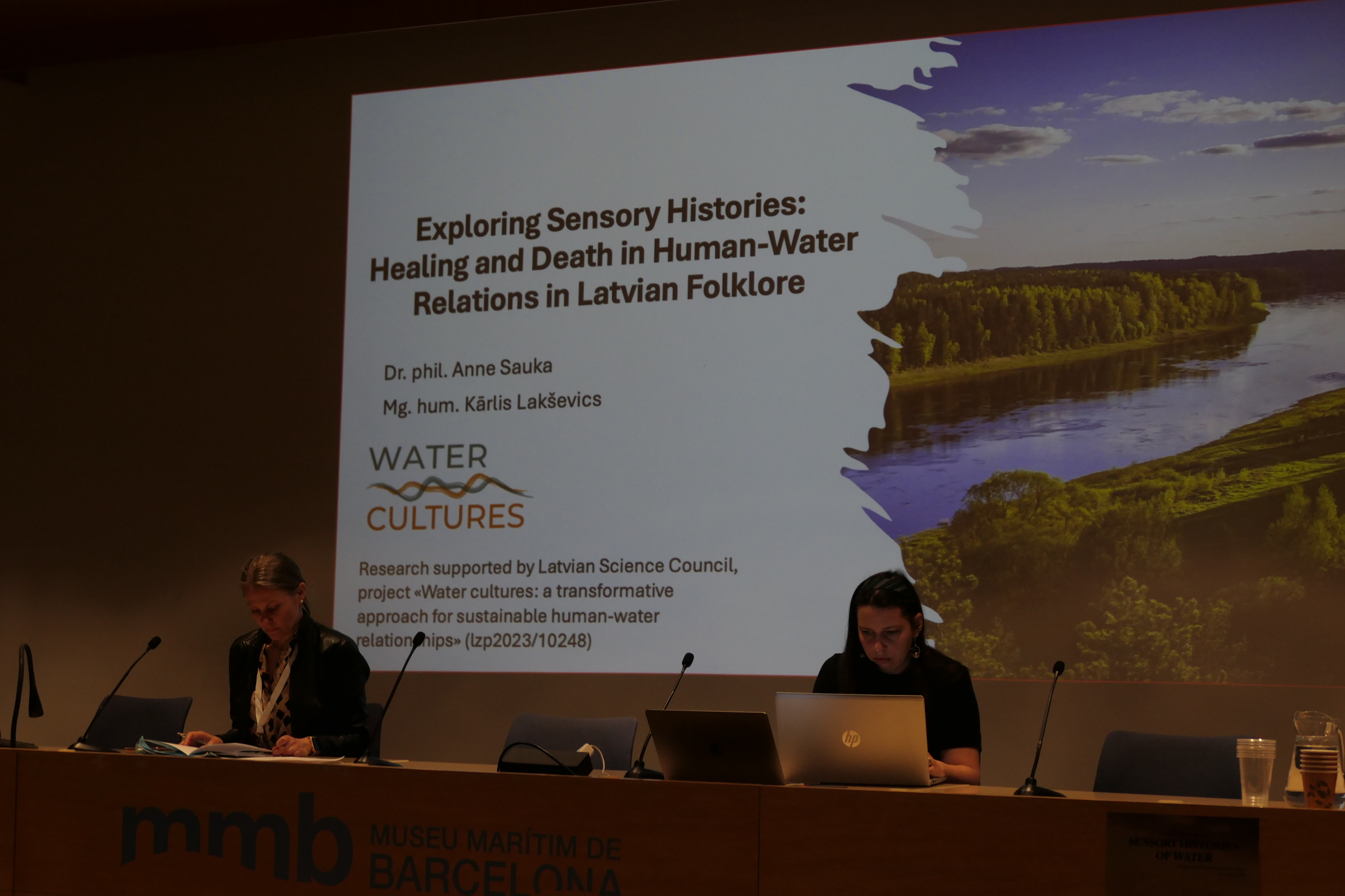 In the foreground is a table with Anne Sauka on the right and another conference participant on the left. In the background is the title slide of the presentation, entitled "Exploring Sensory Histories: Healing and Death in Human-Water Relations in Latvian Folklore".