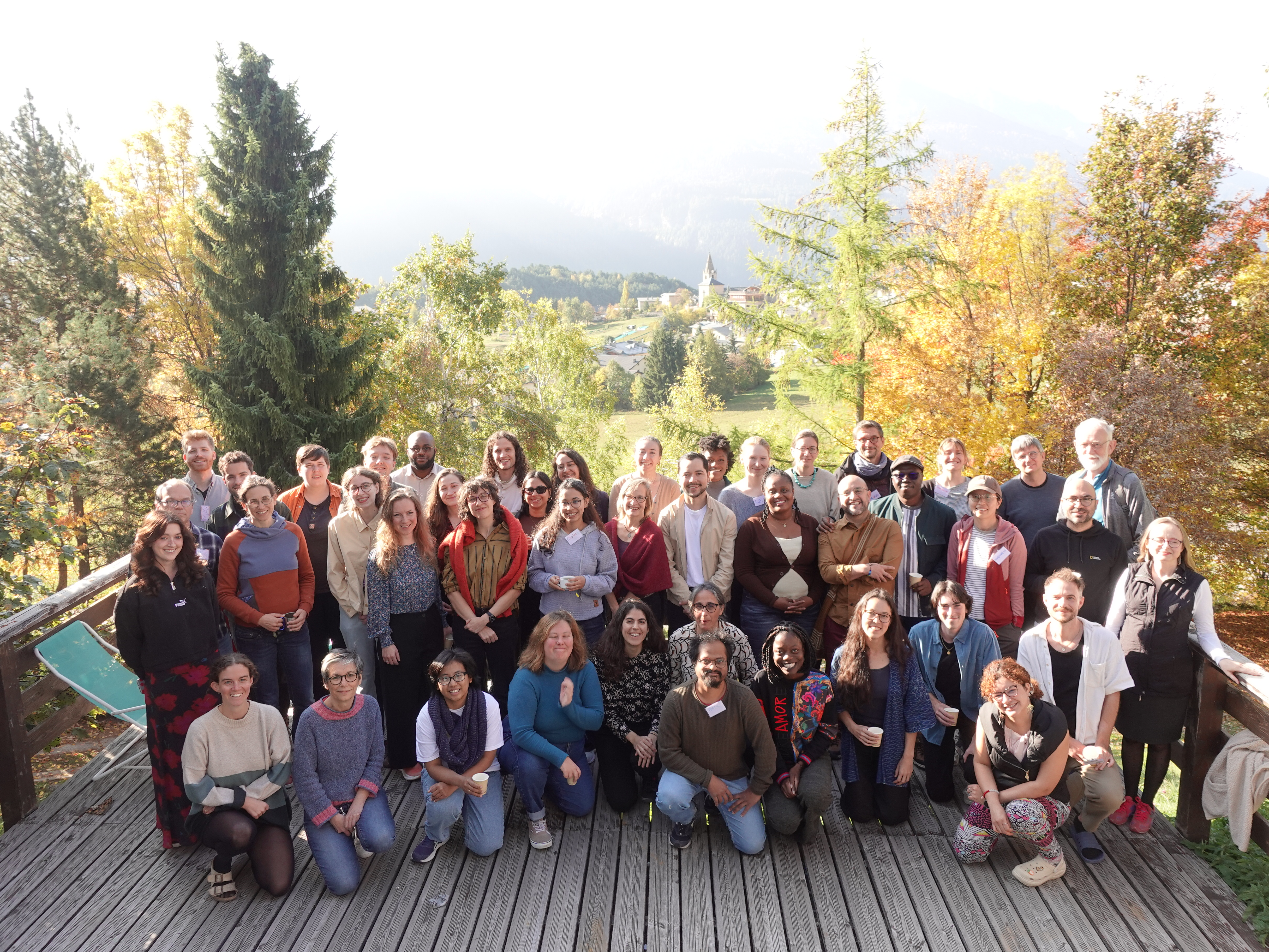 The picture shows a group of more than 20 people sitting on a wooden terrace. Various trees can be seen in the background.