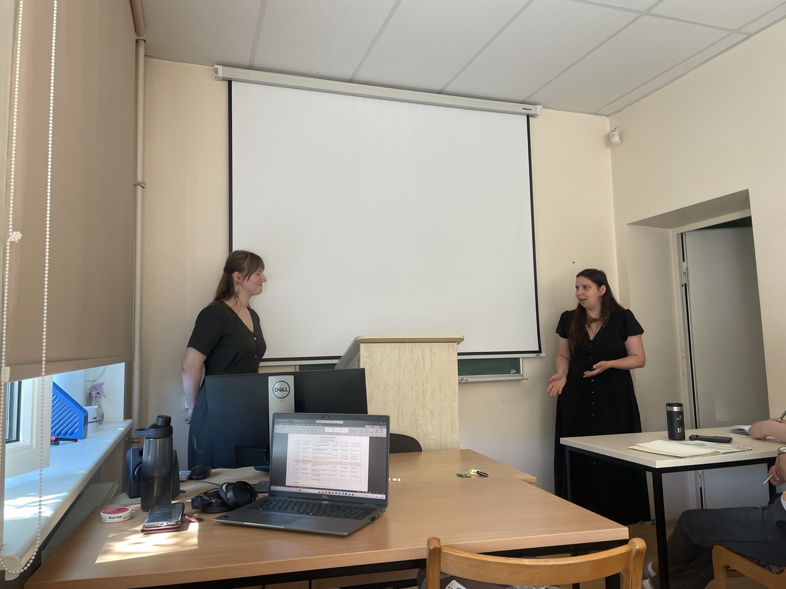 Paula Lūcija Lejiņa stands beside the lectern in a classroom while Anne Sauka addresses the audience. A projection screen is visible in the background, with desks, laptops, and workshop materials in the foreground.
