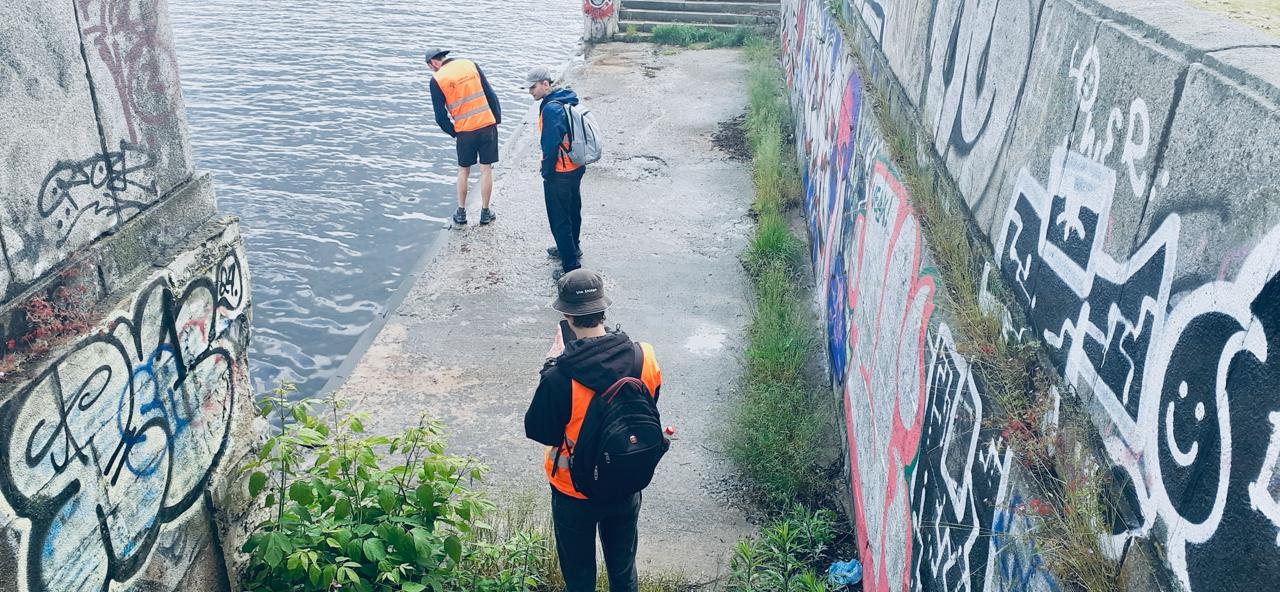 The picture shows three students standing on the concrete edge of the water body. The concrete walls are marked with graffiti drawings in various colors.