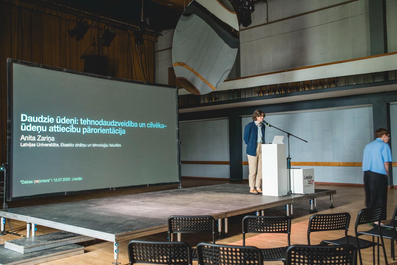 On the left side of the image is a presentation screen with the title "Many Waters: Technological Diversity and the Reorientation of Human-Water Relations." To the right of it, on a raised platform by the microphone, is project researcher Anita Zariņa.
