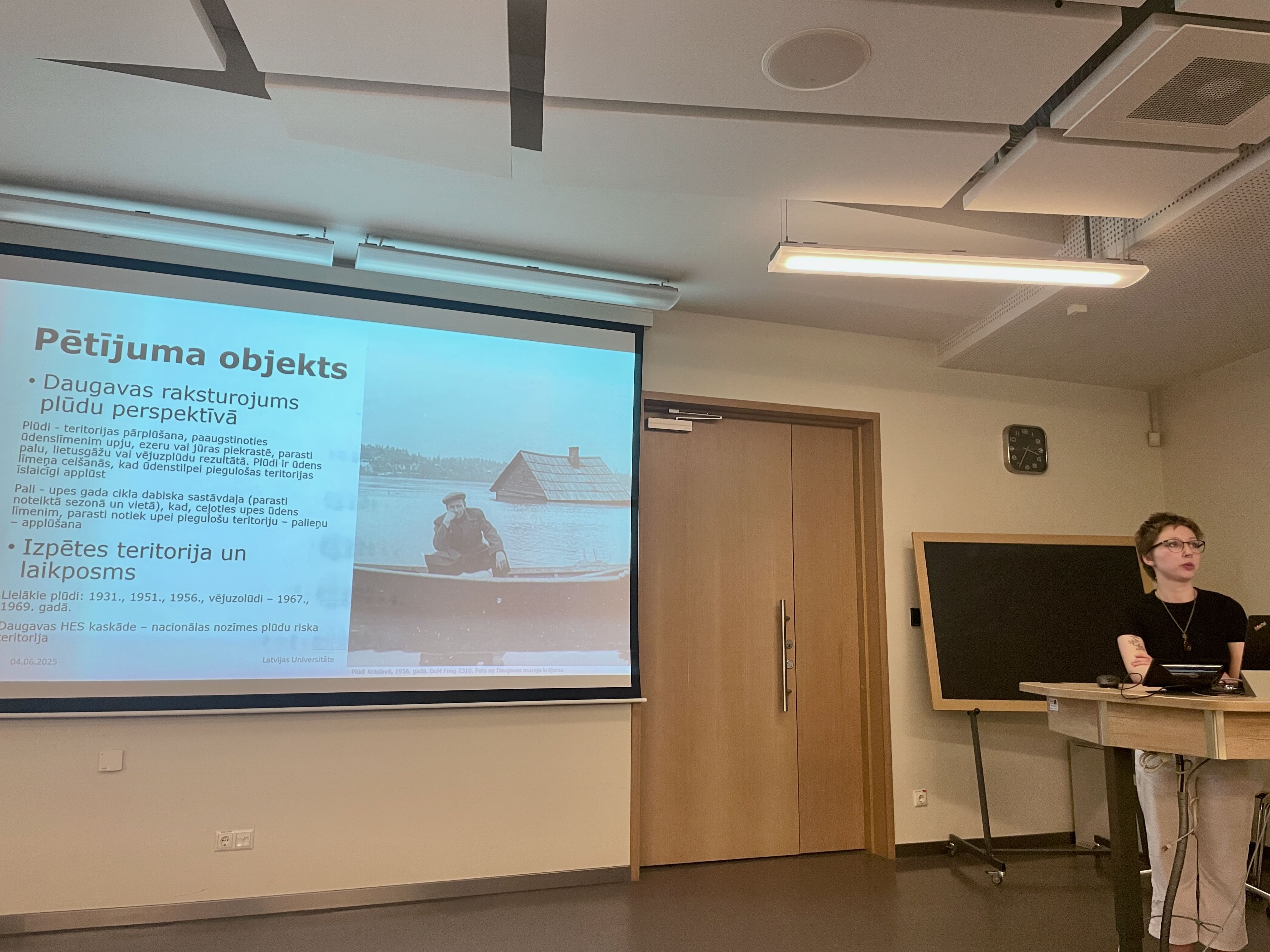 On the right side of the image is presenter Lība Karlīna Varapoga. On the left is the presentation screen with the title "Research Object". On the right side of the presentation screen is an image of a man in a boat with a house in the background that has been flooded, with the water level reaching the roof.