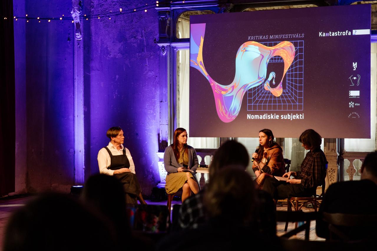 In the central part of the picture you can see the participants of the discussion, from left - Dace Bula, Ieva Astahovska, Anne Sauka, Anita Zariņa. The upper right part of the image shows the presentation screen with the captions "Criticism Mini-Festival. Nomadic Subjects. Cantastrophe". In the foreground of the picture you can see some of the audience.