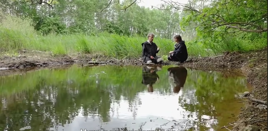 The image shows Māris Olte (left) and Anita Zariņa (right) sitting on a beaver dam while engaged in conversation. In the foreground, the calm water reflects both the speakers and the surrounding landscape. Reeds and trees form the background, creating a natural riverside setting. The scene is filmed from a low angle.