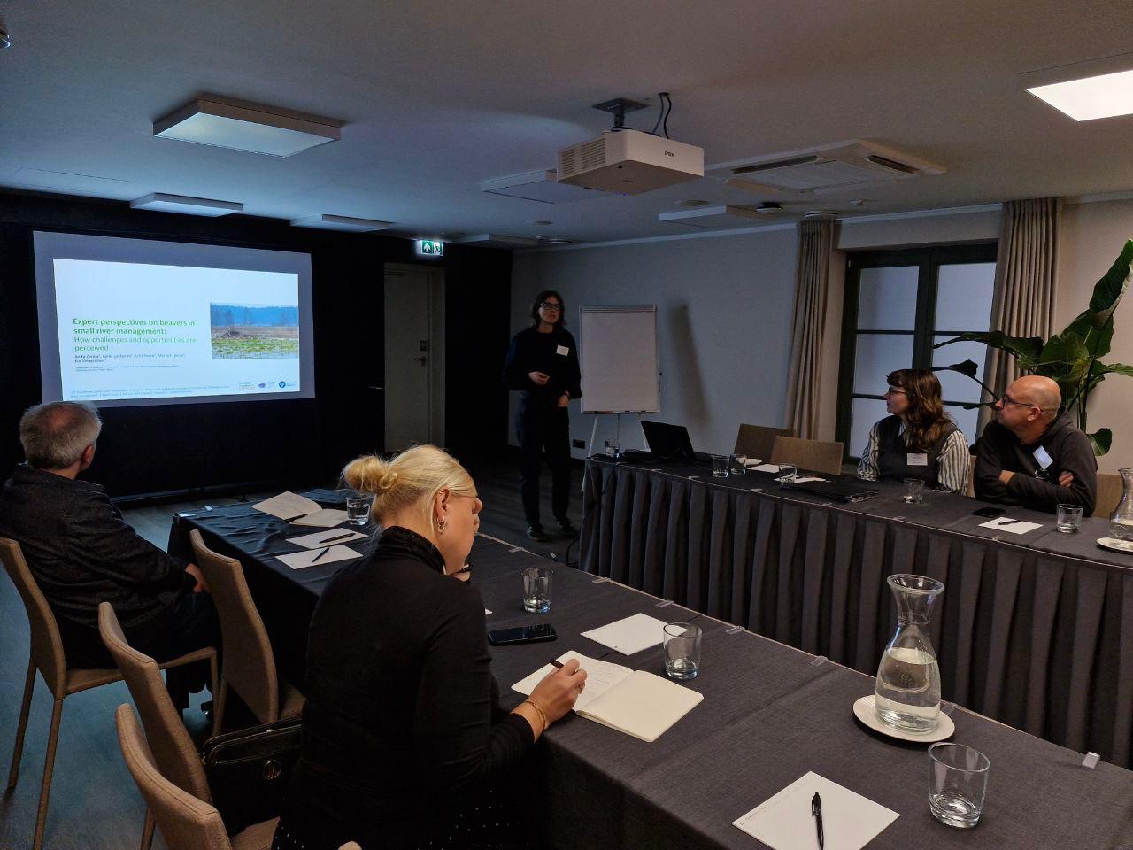 The image shows a presentation of a paper. On the left side, there is a presentation slide with text. In the foreground and on the right side, there is a table with people sitting around it. In the center of the image, in the far distance, there is project researcher Anita Zariņa.