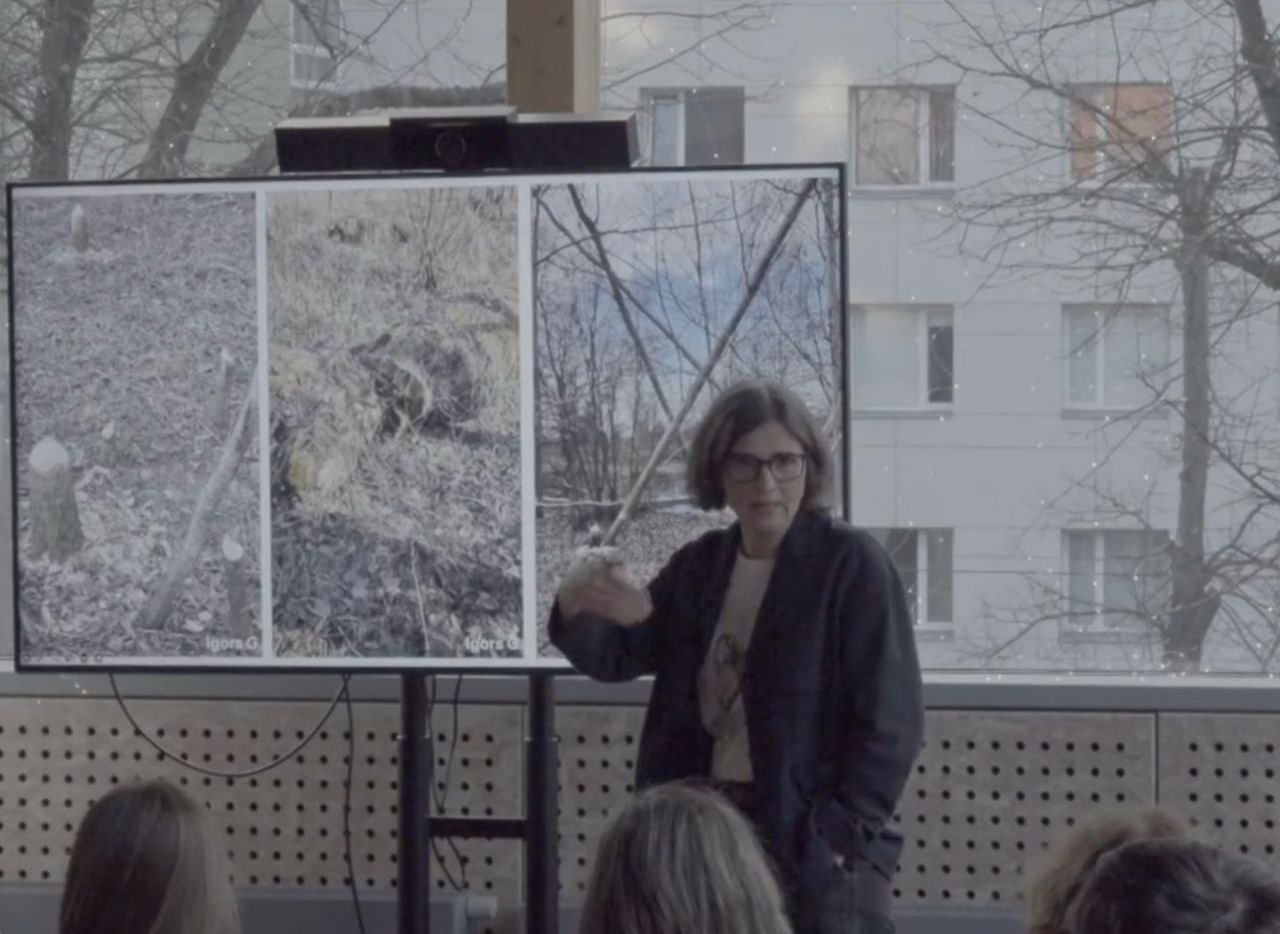 Anita Zariņa is giving a lecture or presentation to an audience. She is standing beside a large display showing several photographs of forested landscapes and natural environments while gesturing as she explains the material. Audience members are visible in the foreground, while large windows in the background overlook trees and a multi-storey building.